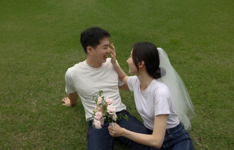흰 티와 청바지 차림으로 잔디에 앉아 신부가 신랑 얼굴을 만지며 장난치는 캔디드 컷이에요.