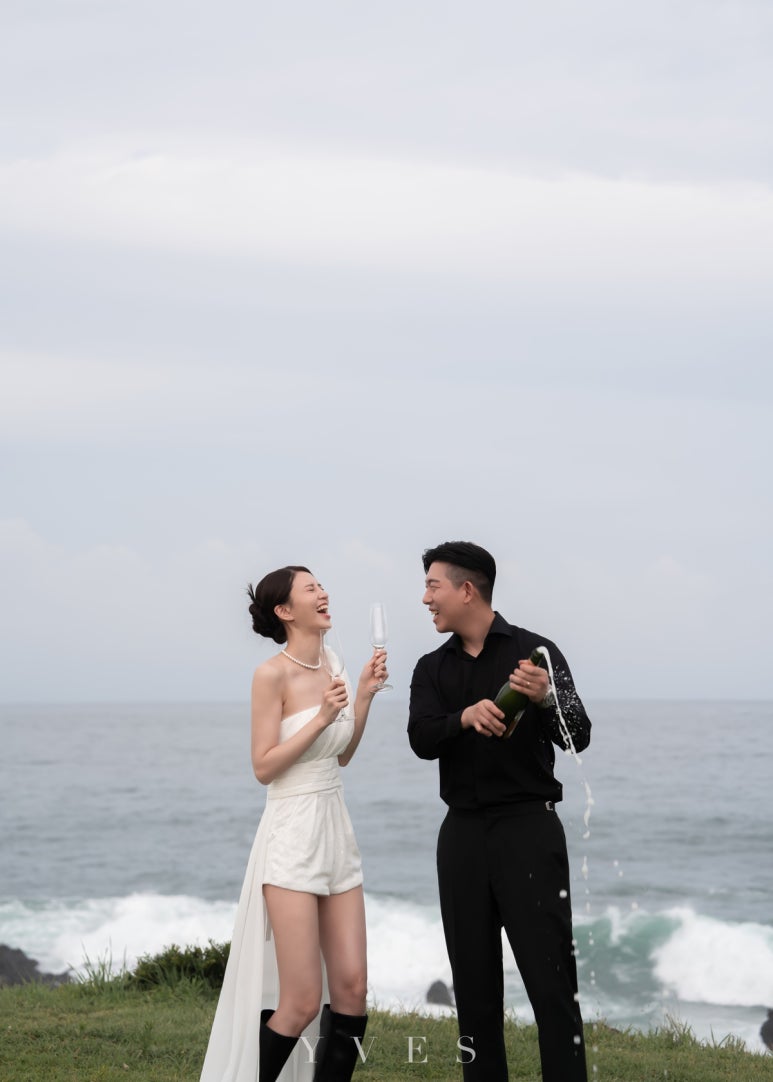 제주 바다 절벽 위에서 신랑이 샴페인을 터뜨리고 미니드레스 신부가 잔을 들고 웃어요
