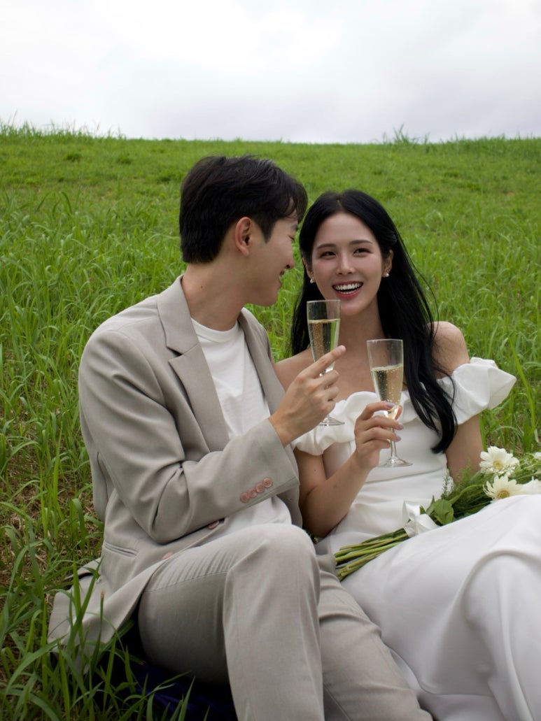 잔디밭에 나란히 앉아 샴페인 잔을 들고 서로 기대어 웃는 컷이에요