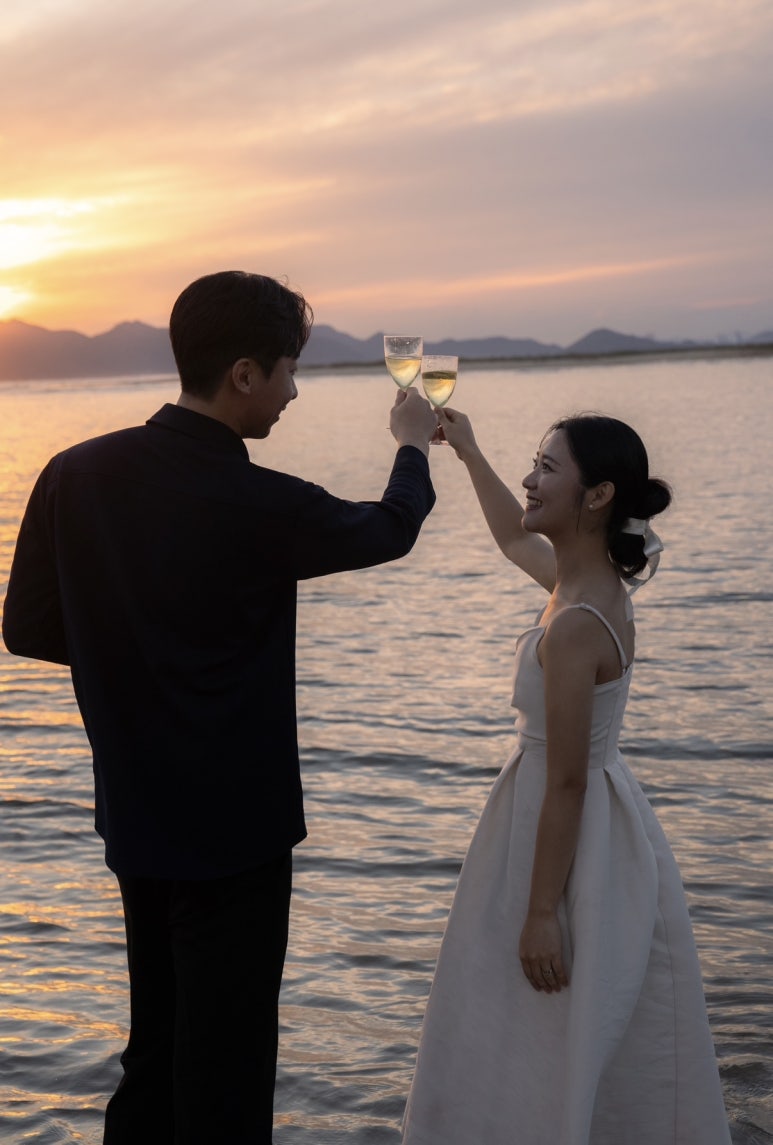 노을 진 바다 앞에서 두 사람이 샴페인 잔을 높이 들어 건배해요