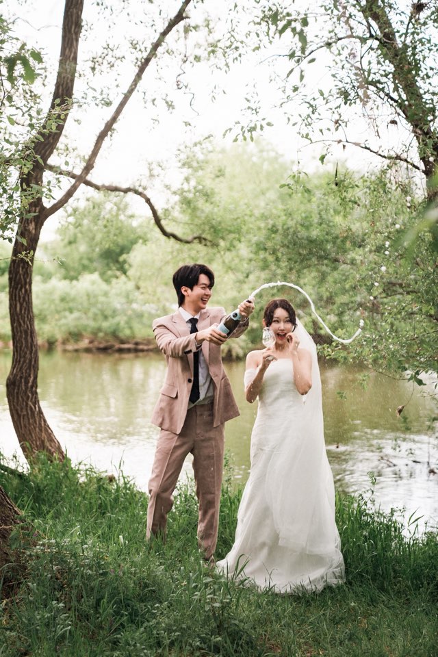 강변 생태공원 나무 사이에서 신랑이 샴페인을 터뜨려 거품이 곡선을 그리고 신부는 놀라 웃어요
