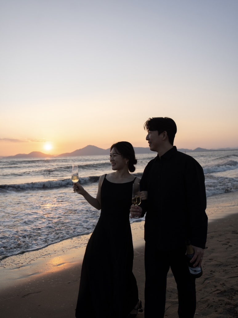 부산 바닷가 노을을 등지고 잔을 든 신부와 병을 든 신랑이 나란히 서 있어요