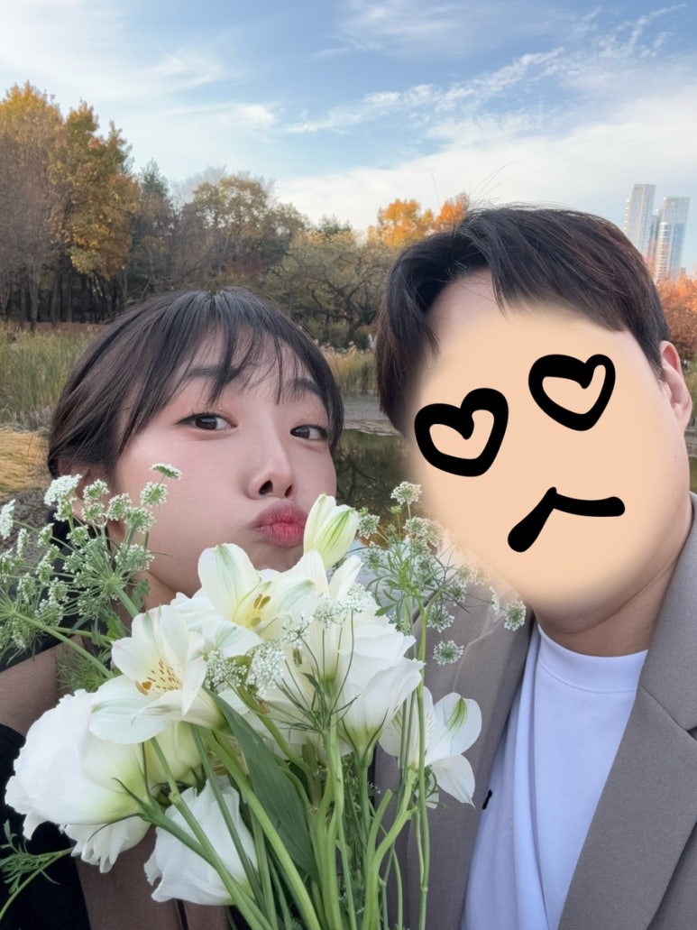 블랙 원피스에 퍼 재킷을 걸친 신부가 흰 꽃다발을 들고 신랑과 휴대폰으로 셀카를 찍는 컷이에요.