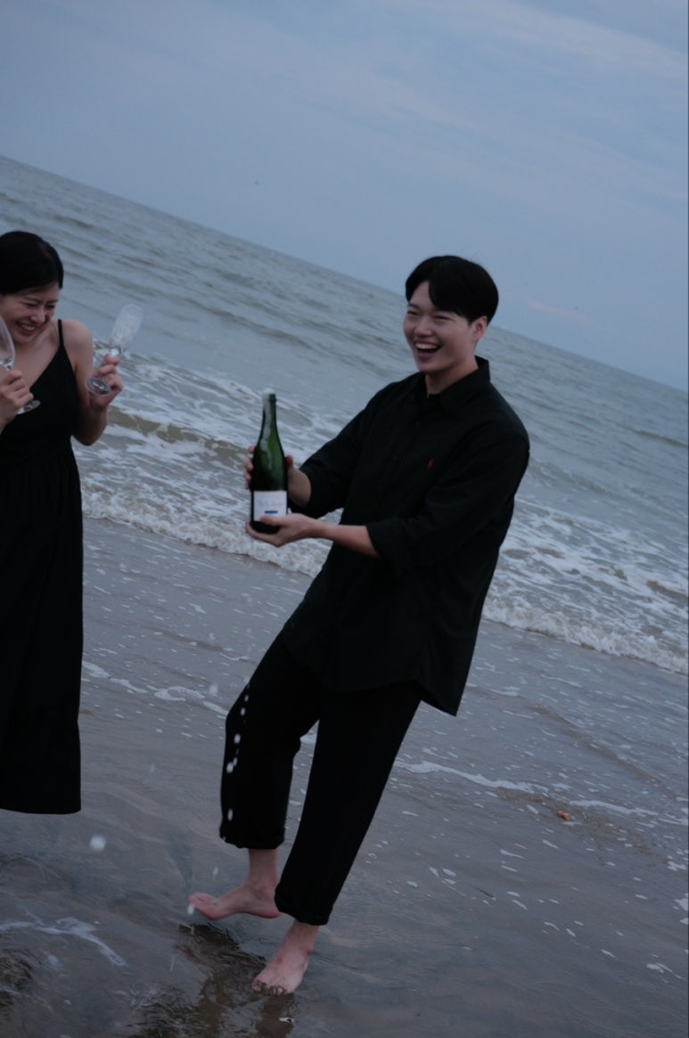 맨발로 바닷물에 선 신랑이 샴페인 병을 흔들며 웃고 신부는 잔을 들고 기다려요