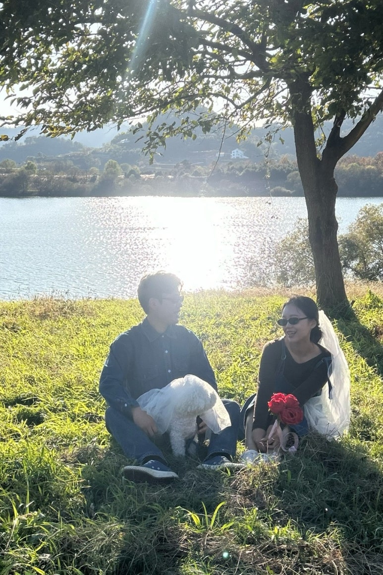 물가 풀밭에 앉아 강아지를 안은 청남방 신랑과 베일에 선글라스를 쓴 신부의 셀프 컷이에요.