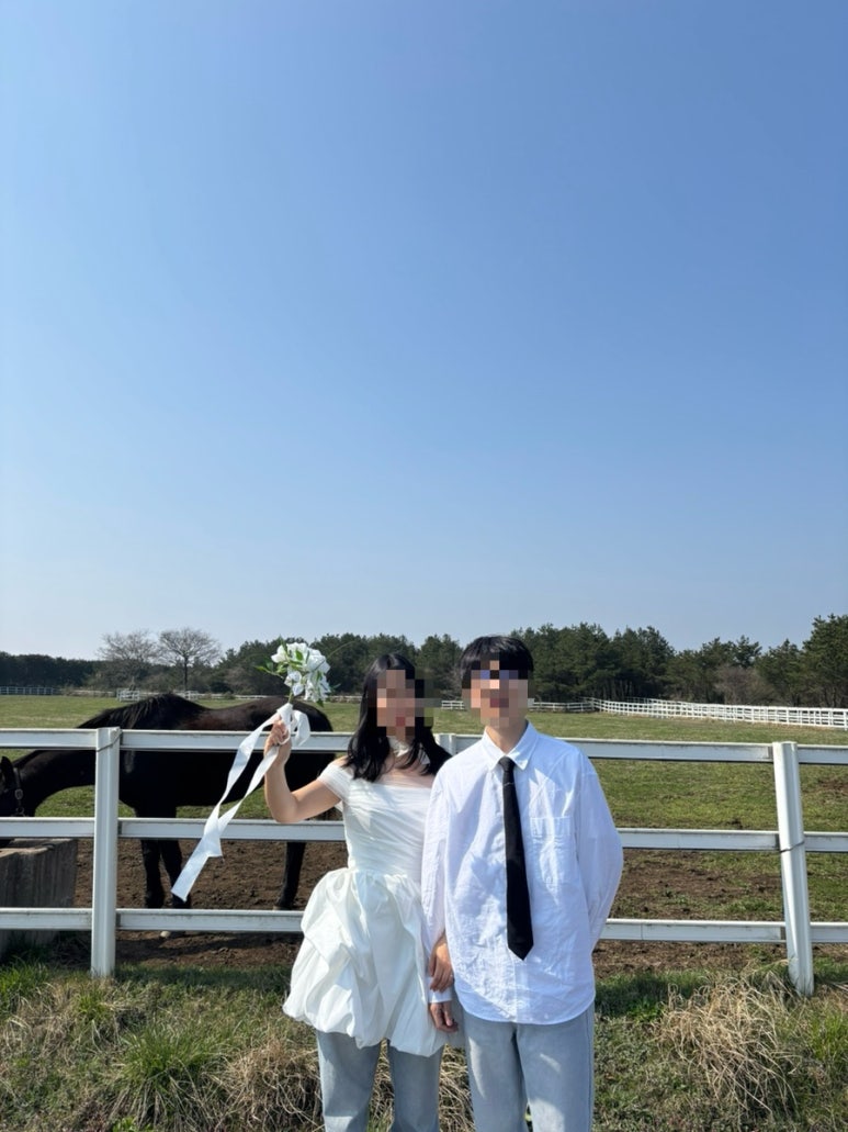 흰 셔츠에 넥타이, 청바지 위 벌룬 미니 원피스로 맞춘 커플이 목장 울타리 앞에서 꽃다발을 흔드는 아이폰 셀프 컷이에요.