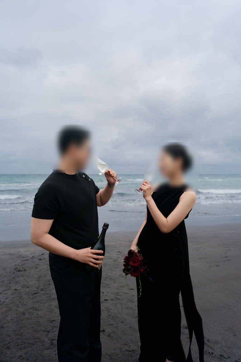 검은 모래 해변에서 신랑은 병을 쥐고 두 사람이 샴페인 잔으로 건배해요