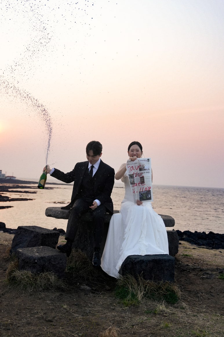 바닷가 돌 벤치에 앉아 신부는 신문 소품을 펼치고 신랑은 샴페인을 옆으로 힘껏 뿜어요