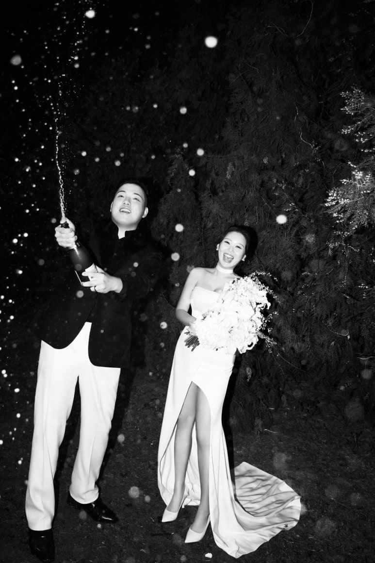 밤 정원에서 흑백으로 담은 샴페인 샤워로 신랑이 병을 위로 쏘고 신부는 부케를 들고 활짝 웃어요