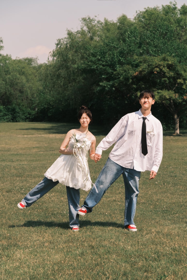 흰 셔츠에 넥타이와 청바지, 청바지 위에 흰 벌룬 미니 원피스를 겹쳐 입은 신부가 손잡고 발을 차올리며 걸어요.