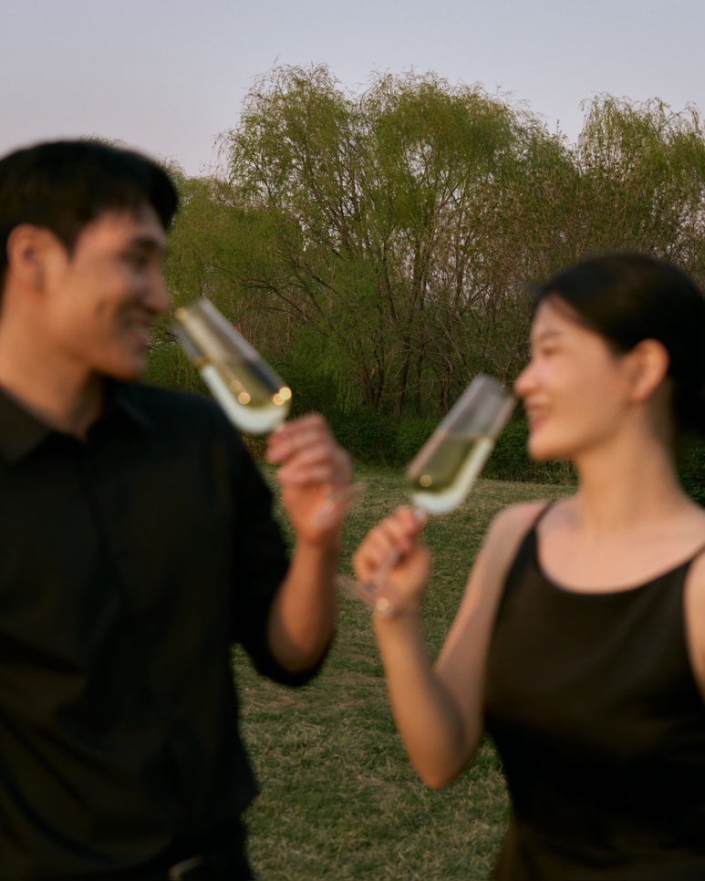 노을공원 풀밭에서 두 사람이 샴페인 잔을 들고 마시며 서로 웃어요