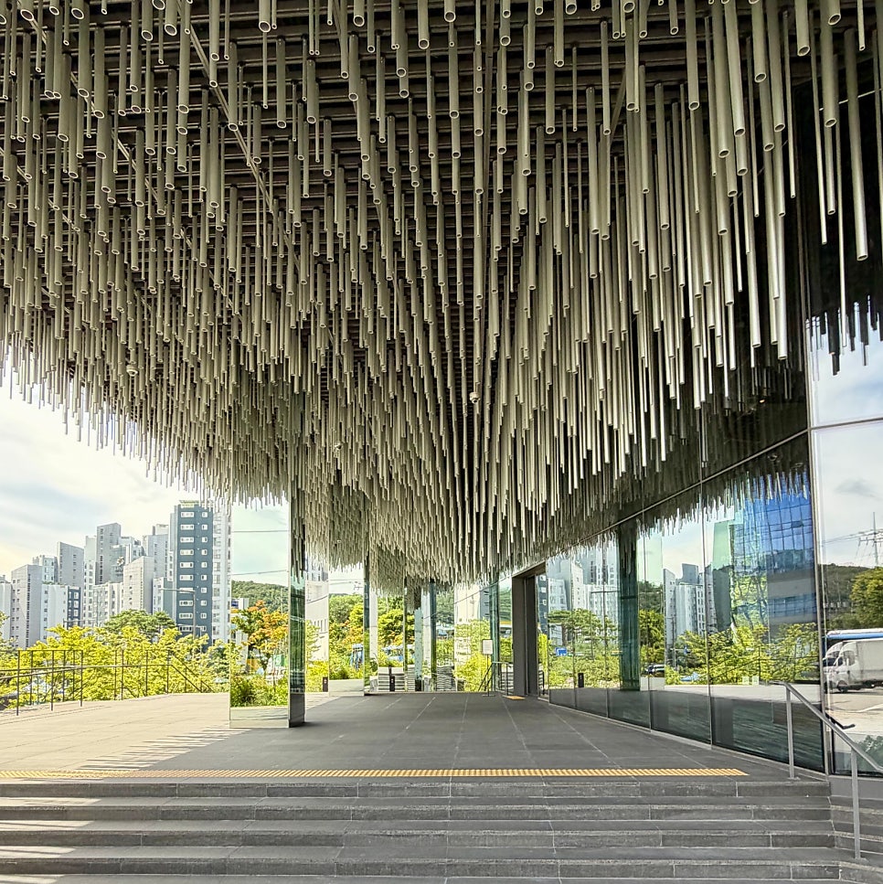 입구 캐노피 아래. 파이프가 천장에서 비처럼 내려오며 도시와 전시 사이의 문턱을 만든다.