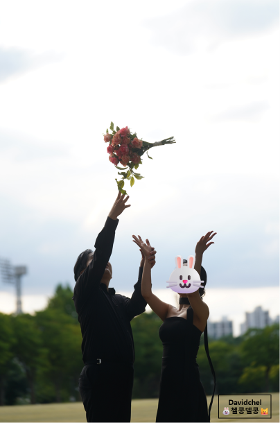 올블랙 커플이 잔디밭에서 빨간 꽃다발을 하늘로 던지는 순간이에요.