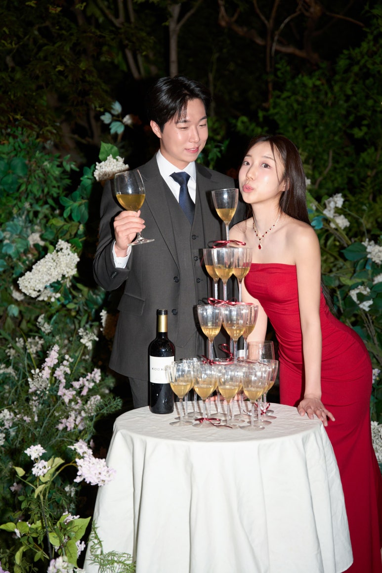 밤 정원의 샴페인 타워 옆에서 빨간 드레스 신부와 신랑이 잔을 들고 장난스럽게 포즈를 취해요