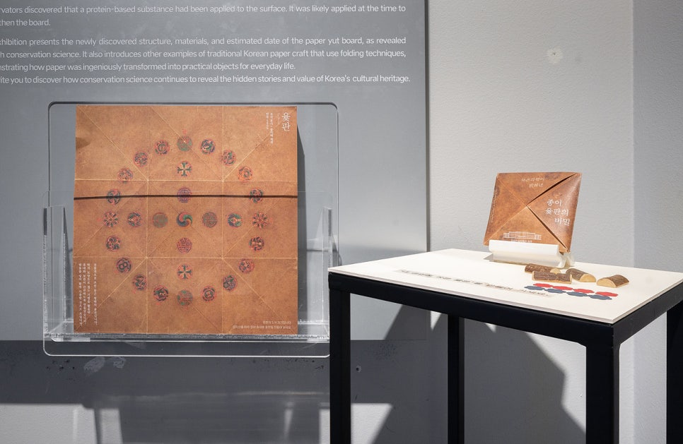 On a table, the leaflet's printed board design beside the folded tile it makes, with small playing sticks and pieces
