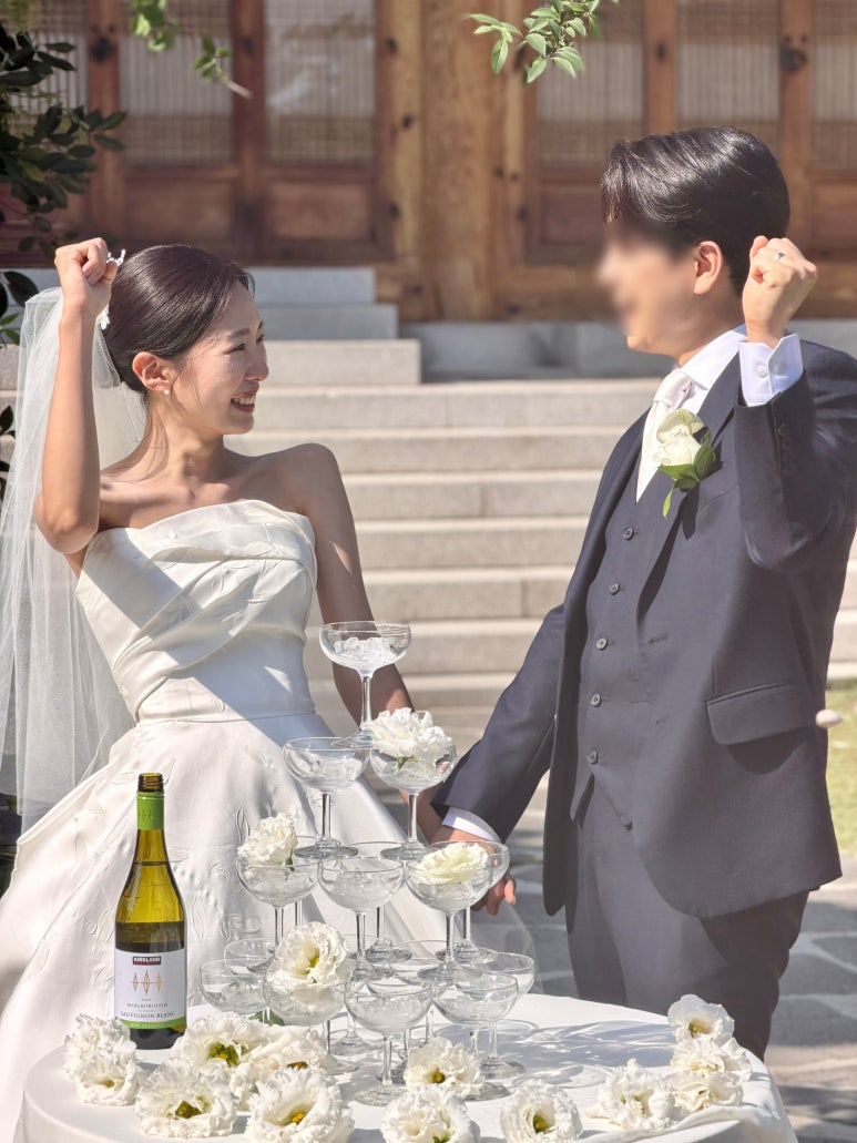 한옥 앞 샴페인 타워 옆에서 두 사람이 주먹을 불끈 쥐며 신나게 환호해요