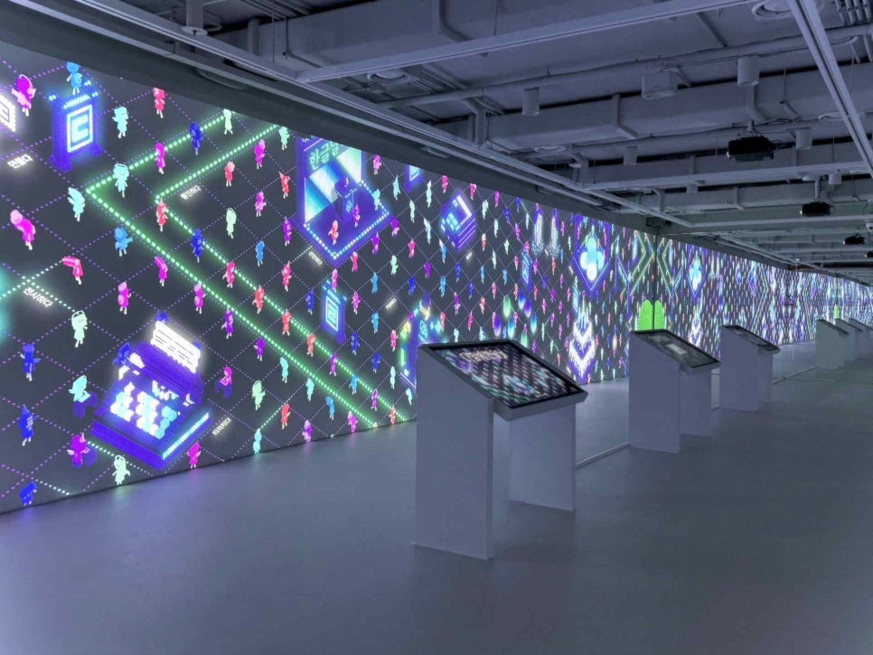 A dark gallery whose entire wall is a digital media screen with Korean letters and sentences flowing across it, a few visitors standing in front