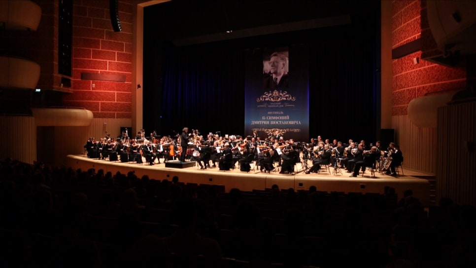 The stage seen from the audience — musicians seated under the lights, with the backs of front-row heads in the foreground