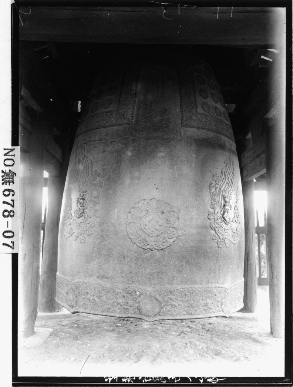 Black-and-white photograph of the bell hanging inside an old roofed pavilion, seen from the front, with the original plate number tag at the bottom
