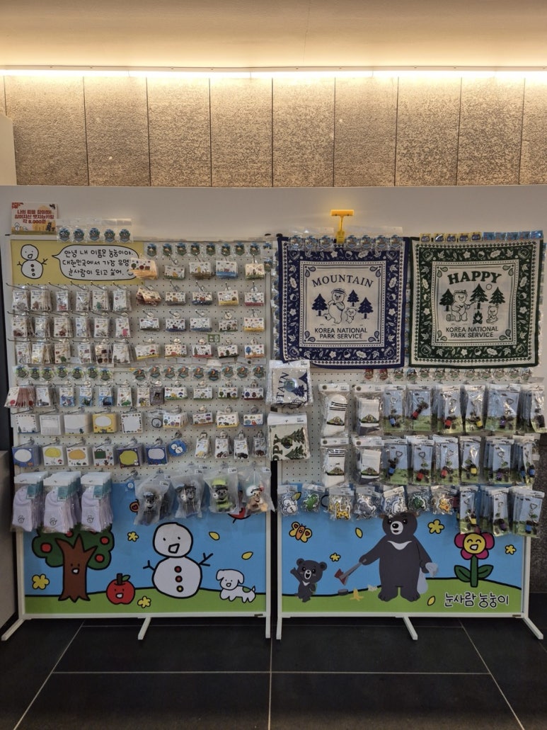 Wooden display shelves inside the National Park Shop at Juwangsan's Sangui Visitor Centre, holding Nungnungi keyrings, badges and fabric goods printed with snowman and Asiatic black bear illustrations