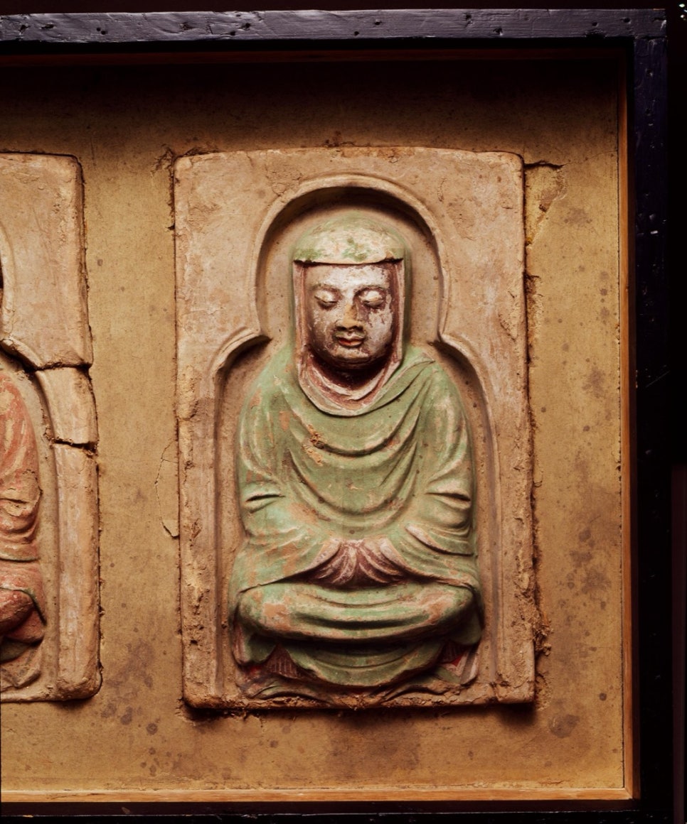 Painted clay panel of a seated monk with eyes closed and hands together inside a niche-shaped frame